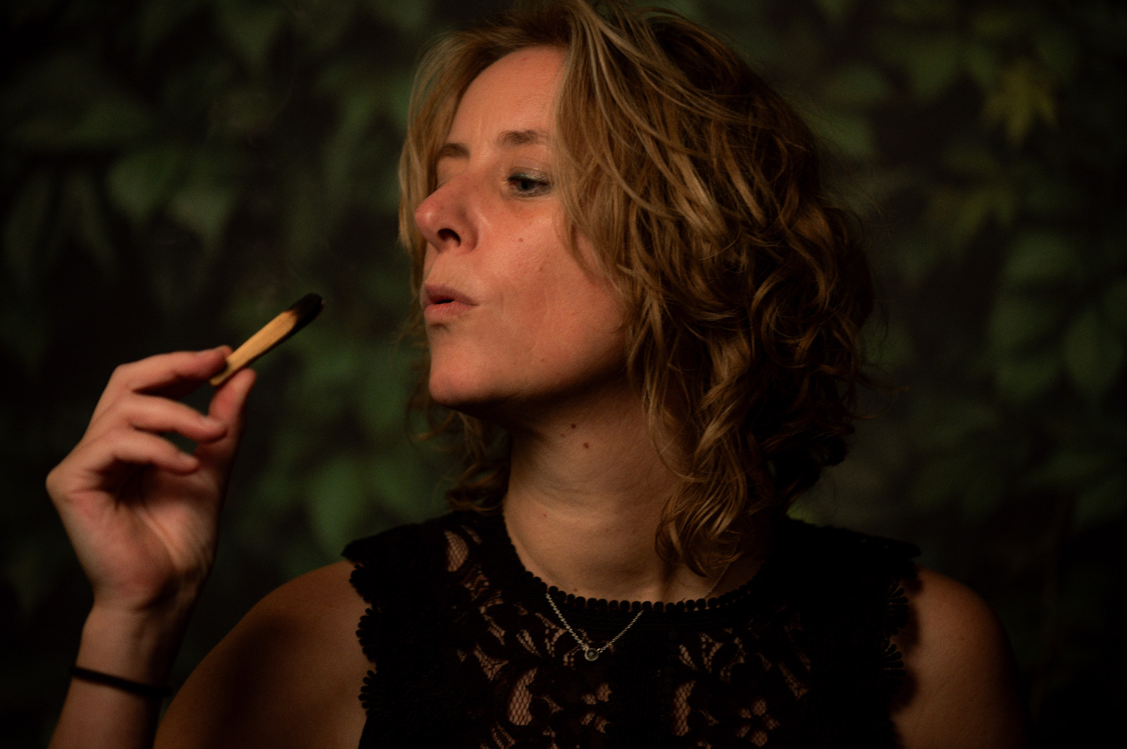 "Woman with curly hair in a black lace top holding a ritual object near her mouth, eyes closed in presence, set against a backdrop of deep green leaves — embodying softness, power, and inner connection."